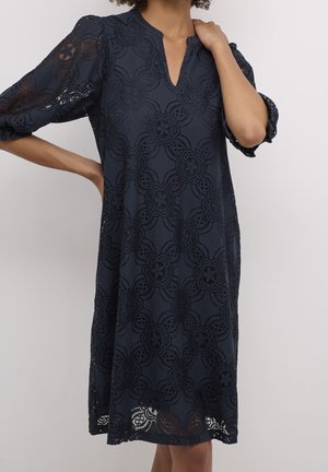 Vrouw draagt een knie-lange marineblauwe jurk met bloemenkantpatroon, V-hals en driekwart doorzichtige mouwen, poserend met één hand aan de nek.
