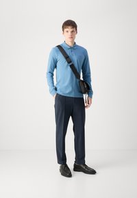 Blaues langärmliges Poloshirt mit Kragen, dunkelblaue maßgeschneiderte Hose und schwarze Schuhe. Eine schwarze Umhängetasche ruht auf der Schulter.