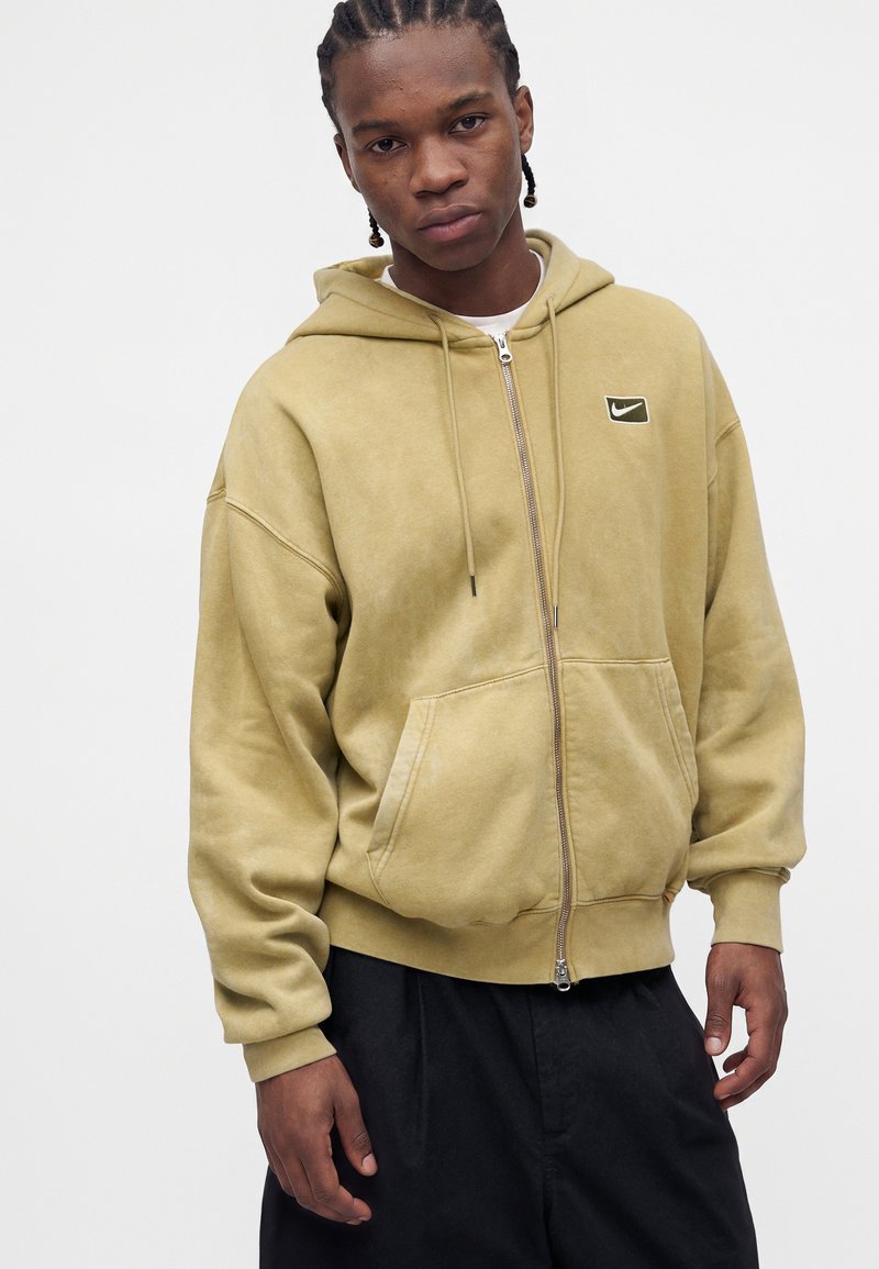 Junger Mann mit geflochtenem Haar, der eine beige Zip-Hoodie mit Nike-Logo und schwarze Hose vor einem schlichten hellen Hintergrund trägt.