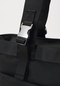 Borsa nera in tessuto con una superficie liscia e testurizzata. Presenta spallacci robusti e una fibbia metallica per una chiusura sicura.