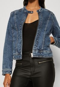 Blå denimjacka med en figurnära design, dragkedjestängning och krage i bandstil. Har framlommer och vita sömnadsdetaljer. Svart topp under.