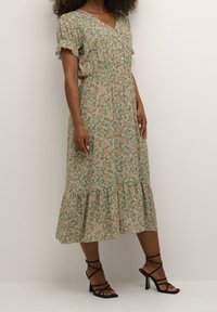 Vihreä kukkamekko, jossa on vaaleanpunaisia ja keltaisia kukkia, V-pääntie, joustava vyötärö ja ryhditetyt lyhyet hihat; yhdistettynä mustiin hihnakenkiin.