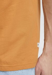 Kortærmet orange t-shirt med en glat tekstur og sidelabel; buet kantdetalje nederst.