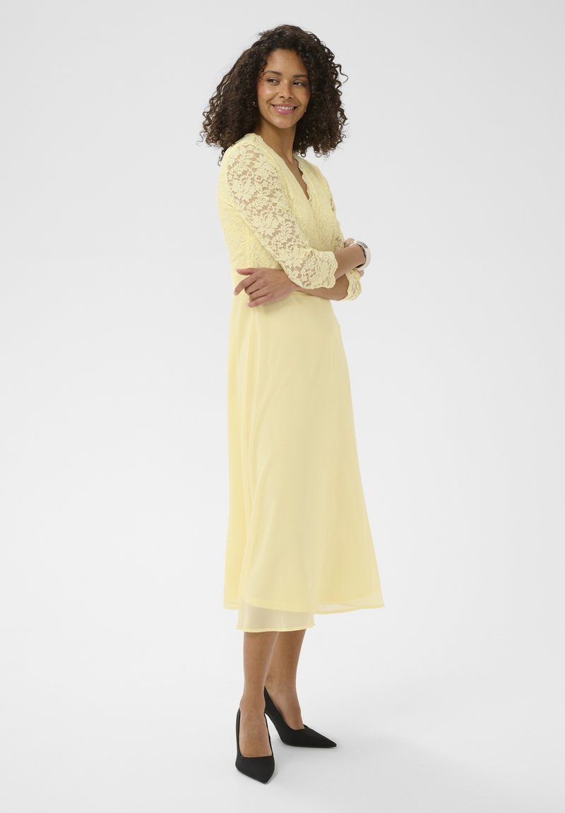 Femme aux cheveux bouclés portant une robe midi jaune à manches en dentelle et des escarpins noirs à bouts pointus, debout les bras croisés, souriante.