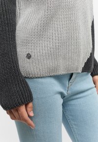 Pull en maille gris et noir avec un motif texturé, arborant un logo rond sur le bord inférieur. Associé à un denim bleu clair.
