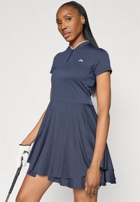 Kobieta w granatowej sukience golfowej z krótkim rękawem trzyma kij golfowy w lewej ręce, na której ma białą rękawiczkę, na jednolitym tle.