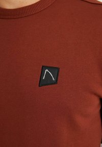Bordová dlouhá košile z hladkého materiálu má černý obdélníkový nášivku s bílým grafickým designem. Kulatý výstřih a žebrovaný lem výstřihu.
