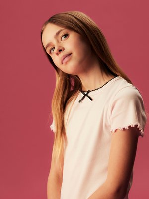 Junges Mädchen mit langen blonden Haaren, das ein hellrosa Hemd mit schwarzem Rand trägt, vor einem einfarbigen rosa Hintergrund steht und leicht nach oben schaut.