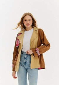 Dvojfarebná bunda s béžovými a hnedými panelmi, károvanými akcentmi na rukávoch, gombíkovými uzávermi a šnúrkami. Nosená cez bielu košeľu.