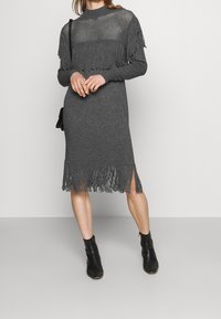 Femme portant une robe en tricot gris foncé avec des détails en frange sur la poitrine, les manches et l'ourlet, associée à des bottes noires à la cheville et un sac à bandoulière.