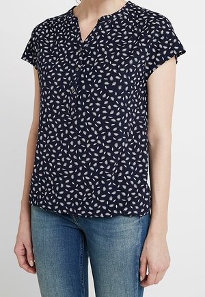 Vrouw die een marineblauwe korte mouw blouse met witte bladpatronen en blauwe spijkerbroek draagt, staand tegen een effen lichte achtergrond.