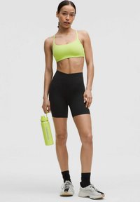 Soutien-gorge de sport vert vif et shorts noirs taille haute. Le mannequin tient une bouteille d'eau vert clair. Les chaussures sont des baskets blanches et grises.