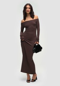 Abito marrone senza spalline, maniche lunghe, design aderente, realizzato in tessuto morbido. La donna tiene in mano una piccola borsa a tracolla nera con texture e indossa sandali neri con tacco.