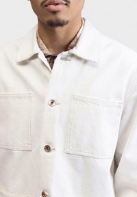 Lys beige knap-jakke med spids krave, to brystlommer og kontrasterende runde knapper. Teksturstof, enkelt design.