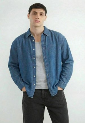 Młody mężczyzna z krótkimi ciemnymi włosami, ubrany w otwartą niebieską koszulę dżinsową na jasnoszarym podkoszulku i czarnych spodniach, ręce w kieszeniach, neutralne tło.