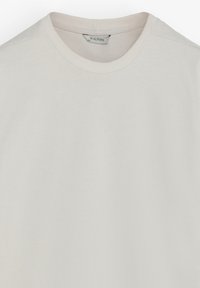 Camiseta de cuello redondo en beige claro, hecha de algodón suave. Presenta una textura lisa sin patrones ni adornos visibles. Etiqueta visible en el cuello.