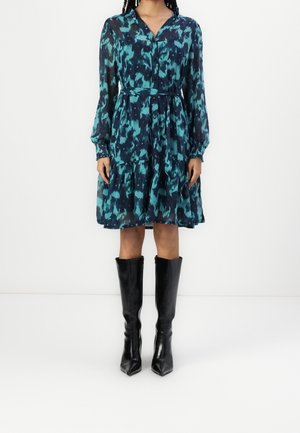 Femme portant une robe à manches longues à imprimé abstrait bleu sarcelle et bleu marine avec une ceinture à la taille, associée à des bottes noires montantes, debout sur un sol blanc.