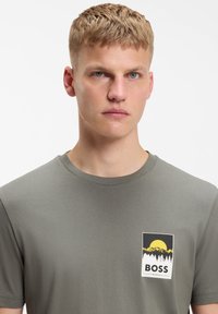 Νέος άνδρας με κοντά ξανθά μαλλιά, ντυμένος με γκρι μπλουζάκι που απεικονίζει ένα κίτρινο ήλιο, βουνό και δέντρο, με το κείμενο «BOSS».