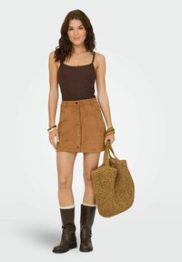 Top sin mangas marrón con tirantes finos, complementado con una minifalda de ante color beis con botones frontales, combinado con botas marrones hasta la rodilla y un bolso de fieltro.