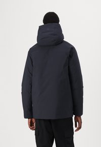 Marineblå dunjakke med en stor, justerbar hætte og sømløst skulderdesign. Har et glat, vandafvisende stof og en tætsiddende pasform.