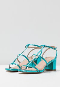 Turkoosi korkokenkät, joissa on metalliset hihnat, avokärkinen muotoilu, neliöalusta ja minimaalinen yksityiskohtaisuus tyylikkään ilmeen saavuttamiseksi.