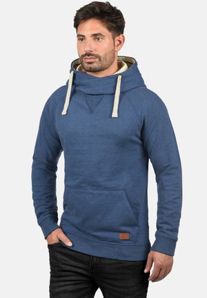 Sudadera azul marino con capucha de doble capa, cordones crema, bolsillo frontal y parche de cuero con el logo en el dobladillo. Textura de tela suave.