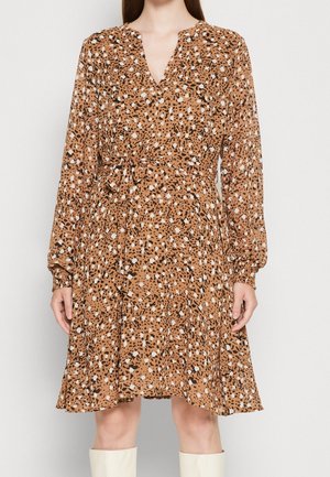 Femme portant une robe marron longueur genoux avec des taches abstraites noires et blanches, manches longues, col en V et ceinture nouée à la taille.