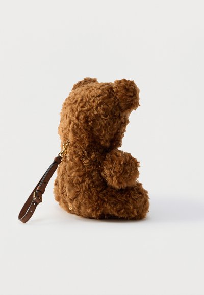 Καφέ τσάντα σε σχήμα αρκουδάκι με κατσαρά μαλλιά, με χρυσό κούμπωμα και προσαρμοζόμενο καφέ δερμάτινο ιμάντα, εμφανιζόμενη από το πλάι σε λευκό φόντο.