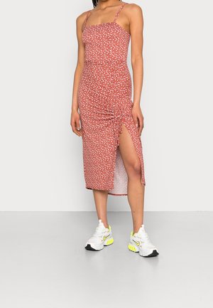 Femme portant une robe midi rouge sans manches à pois blancs avec une fente latérale, associée à des baskets blanches et jaune néon.