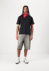 Tricou negru cu mânecă scurtă, pantaloni scurți din denim gri, batic roșu, adidași albi cu accente negre, stând în fața unui fundal alb.