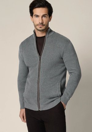 Mężczyzna ubrany w szary sweter na zamek z wysokim kołnierzem i czarne spodnie, stojący na jednolitym beżowym tle.