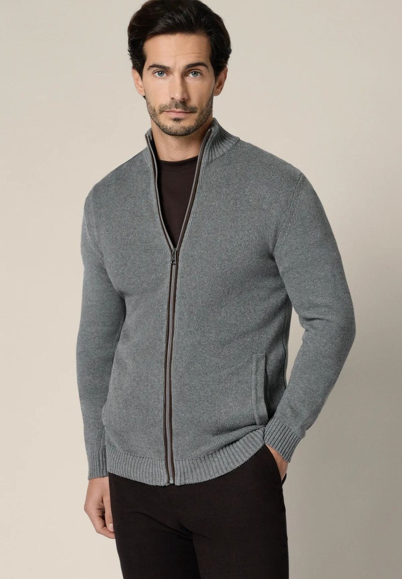 Mężczyzna ubrany w szary sweter na zamek z wysokim kołnierzem i czarne spodnie, stojący na jednolitym beżowym tle.