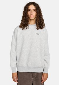 Sudadera gris claro hecha de tela suave, con cuello redondo, puños acanalados y un pequeño logo negro en el pecho.