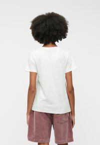 Fehér pamut póló rövid ujjakkal, laza szabású, kerek nyakkivágással, vörös bordás shorttal viselve. A háttér sima fehér.