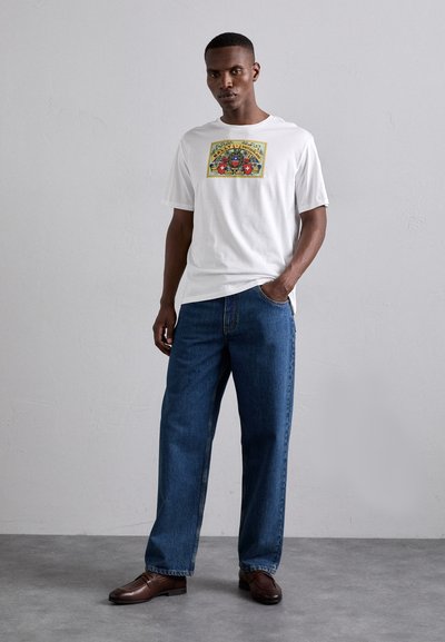 Camiseta de algodón blanca con un colorido estampado gráfico en la parte frontal, combinada con jeans de mezclilla azul de corte holgado y zapatos de cuero marrón.