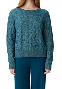 Frau, die einen türkisfarbenen, kabelgestrickten Pullover mit geripptem Saum und Bündchen trägt, kombiniert mit passenden türkisfarbenen Hosen, von Hals bis Hüfte gezeigt.