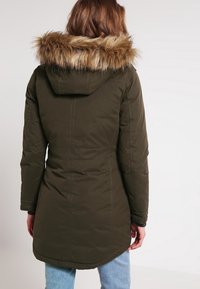 Woman wearing a dark green parka coat with a fur-lined hood, viewed from the back, standing against a plain light background.