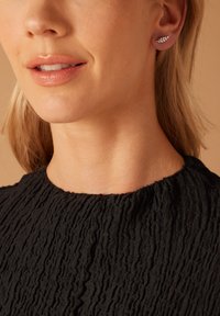 Silberne blattförmige Steckerohrringe an einem Model, kombiniert mit einem strukturierten schwarzen Oberteil. Sanfter Hautton, neutraler Hintergrund.