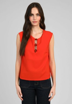 Femme aux longs cheveux bruns porte un haut sans manches rouge vif avec trois boutons décoratifs et un jean en denim foncé, debout devant un fond gris.