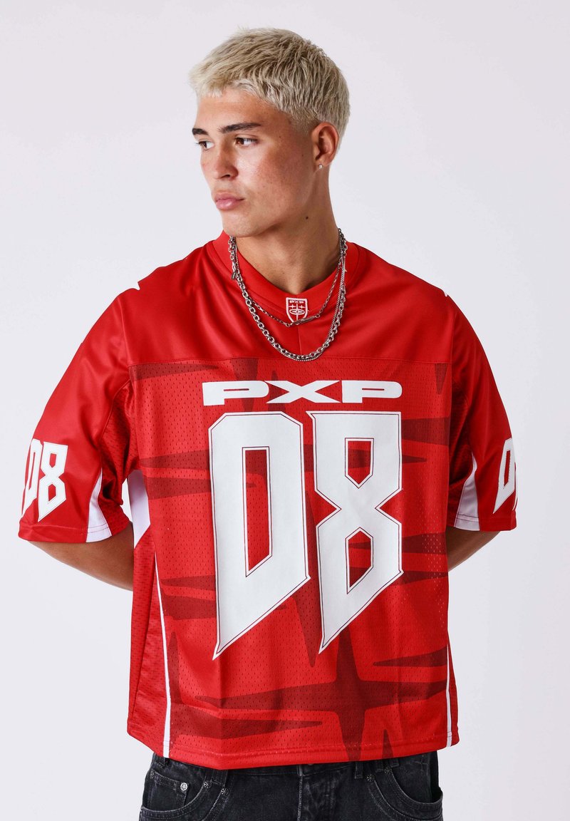 Jeune homme aux cheveux blonds courts portant un maillot de sport rouge avec les logos blancs "08" et "PXP", regardant sur le côté les mains derrière le dos.