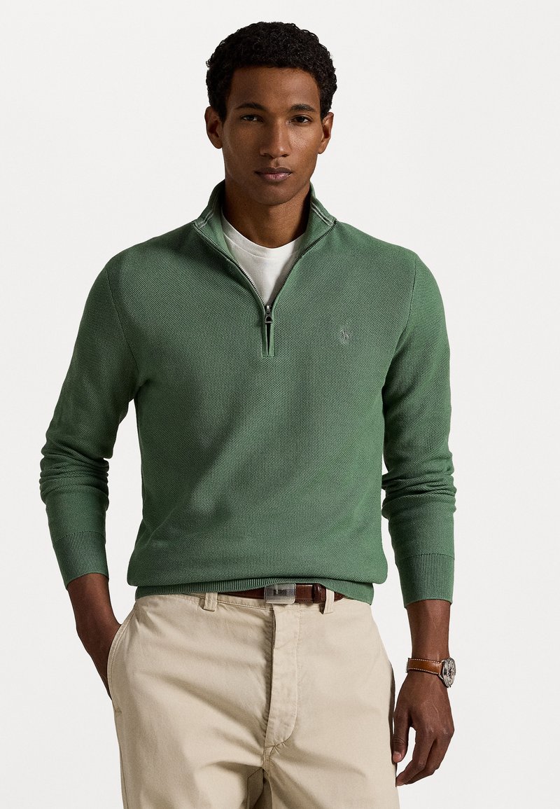 Green pullover sweater with a zip collar, knitted texture, ribbed cuffs, and a small logo on the chest. Worn over a white t-shirt and beige pants.