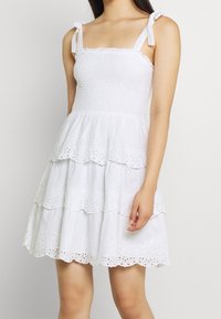 Žena v bílé šaty s vrstvami a dírkovaným vzorem, s uvázanými ramínky a nařaseným živůtkem, stojící na jednobarevném pozadí.