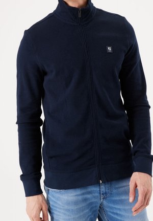 Navy blue zip-up jacket with a high collar, ribbed texture, long sleeves, and a small logo patch on the left chest.