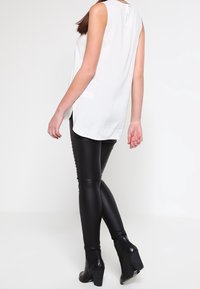 Haut blanc sans manches avec un détail de bouton au dos, associé à un pantalon noir ajusté et des bottines à talons, vu de dos.