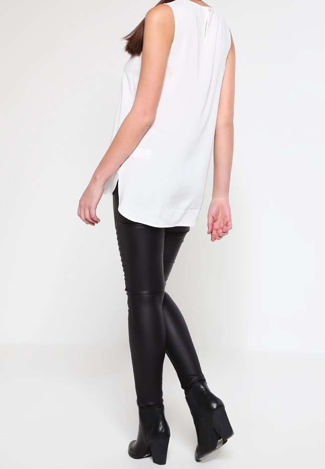 Haut blanc sans manches avec un détail de bouton au dos, associé à un pantalon noir ajusté et des bottines à talons, vu de dos.