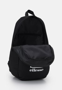 Sac à dos noir en tissu, avec une forme arrondie, une poignée supérieure, une fermeture éclair et une impression de logo à l'avant. L'intérieur dispose d'une poche zippée.