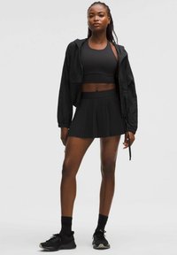 Crni atletski outfit koji se sastoji od uskog crop topa i plisirane suknje. Model nosi laganu crnu jaknu i crne tenisice.