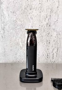 Tondeuse électrique BaByliss noire et argent sur base de chargement, posée sur une surface grise avec un mur blanc texturé en arrière-plan et un protège-tondeuse noir à proximité.