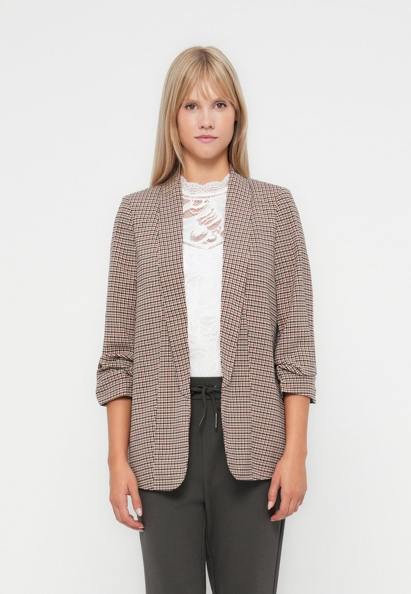 Blazer à carreaux en brun, noir et blanc. Présente une coupe ajustée, un devant ouvert et des manches trois-quarts. Porté sur un top en dentelle blanche.