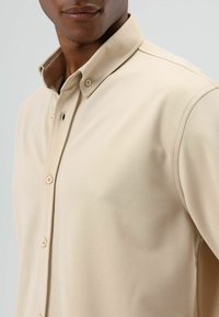 Beige overhemd met knopen, voorzien van een klassieke kraag, een gladde textuur en subtiele stikdetails. De knopen aan de voorkant zijn zichtbaar.